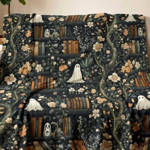 May include: A dark-colored throw blanket featuring a repeating pattern of bookshelves filled with books, interspersed with white ghost figures, winding tree branches, and clusters of light-colored flowers in shades of cream and soft orange.