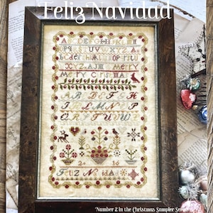 May include: A framed cross-stitch sampler featuring the alphabet, the text "Merry Christmas" and "Feliz Navidad", and motifs of birds, hearts, and floral baskets. The sampler is enclosed in a dark brown wooden frame with a textured, mottled finish.