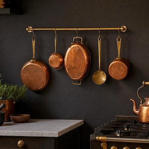 Peut inclure: Batterie de cuisine en cuivre suspendue à une barre en laiton contre un mur sombre. L'ensemble comprend une grande poêle, une petite casserole, un plat ovale, une cuillère ajourée et une petite marmite. La cuisine comprend également un plan de travail en marbre et une cuisinière à gaz.
