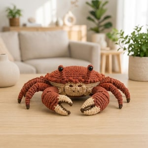 May include: A crocheted crab plush toy featuring a textured rust-coloured shell, a light beige underside, and two black bead eyes. The crab has eight articulated legs and two large front claws with beige tips, all crafted with detailed yarn stitching.