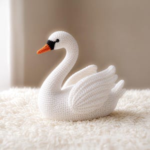 Könnte beinhalten: Ein handgehäkelter weißer Schwan mit langem, gebogenem Hals und ausgebreiteten Flügeln. Der Schwan hat eine schwarze Schnabelspitze, einen orangefarbenen Schnabel und ein kleines schwarzes Auge. Der Schwan sitzt auf einer flauschigen weißen Oberfläche.