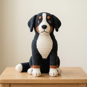 Puede incluir: Un peluche de ganchillo que representa un perro de montaña bernés. El perro es principalmente negro con marcas blancas y marrones, incluyendo un pecho y una cara blancos. Está sentado en una mesa de madera, con un fondo neutro.
