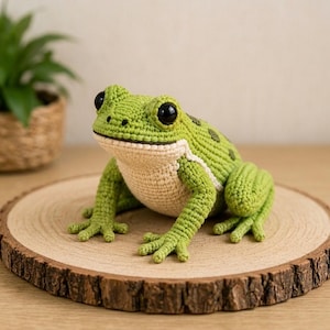 Op de afbeelding: Een handgemaakt gehaakt kikkerfiguurtje met een felgroen getextureerd lichaam met donkergroene vlekken op de rug en een crèmekleurige buik. De kikker heeft grote, ronde, glanzend zwarte ogen en gedetailleerde zwemvliezen aan de voeten.
