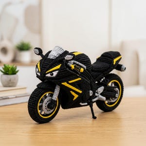 Op de afbeelding: Een handgemaakt gehaakt motorspeelgoed in zwart met levendige gele accenten en lichtgrijze details op de wielen, het stuur en de voorruit. Het sportmotorontwerp heeft een getextureerde garen afwerking, een standaard en een gedetailleerde voorzijde.
