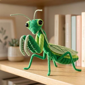 May include: A handcrafted crochet praying mantis toy featuring a vibrant green body, textured yarn stitching, two long antennae, and large black plastic eyes. The insect figure has articulated front raptorial legs and detailed wings.