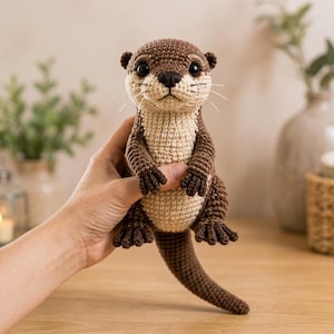 Può includere: Una piccola lontra all'uncinetto fatta a mano con un corpo marrone scuro e petto e pancia beige chiaro. Il peluche presenta grandi occhi neri, un naso ricamato in marrone scuro e sottili baffi bianchi che sporgono dal muso.