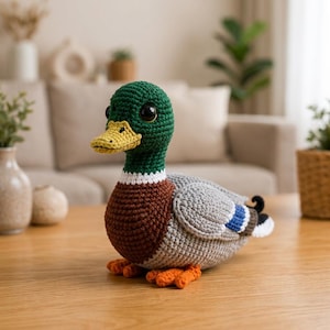 May include: A crocheted mallard duck plush toy featuring a dark green head, a yellow beak, a white neck ring, a brown chest, and a grey body with blue and black wing accents. The duck has large black eyes and orange webbed feet.