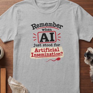 May include: A light gray short-sleeved t-shirt featuring the black and red text "Remember when AI just stood for Artificial Insemination?" with a red rectangular border around "AI" and a small red sperm icon below the text.