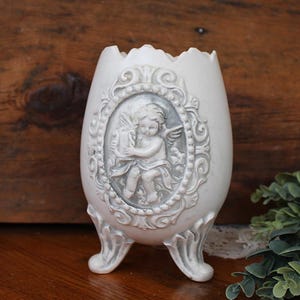 May include: An egg-shaped white vessel with a jagged top edge, supported by three ornate feet. The front features a raised oval medallion depicting a cherub playing a harp, framed by intricate scrollwork and bead detailing in a grey wash.