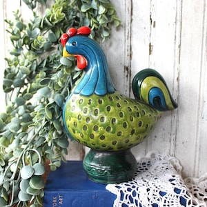 May include: A ceramic rooster figurine featuring a blue neck, a bright red comb and wattle, and a yellow beak. The body is painted in shades of green with dark circular spots, while the tail feathers display blue, green, and yellow accents.