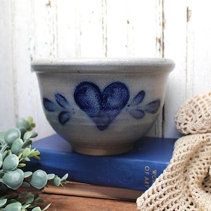 May include: A speckled light gray ceramic bowl with a wide rim and a tapered base. The exterior features a hand-painted dark blue heart design centered on the front, flanked by two sets of three small blue leaf-like brush strokes on each side.