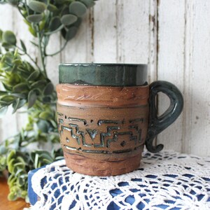 May include: A ceramic mug with a textured, unglazed brown clay body featuring a dark green glazed rim and handle. The exterior surface displays a carved geometric pattern filled with dark green glaze, contrasting with the rough, earthy texture.