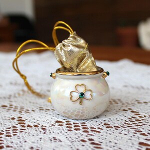 May include: A small, iridescent white ceramic cauldron featuring a gold-toned shamrock emblem with a central green rhinestone. A metallic gold fabric pouch with a gold drawstring cord is tucked inside the opening of the rounded vessel.