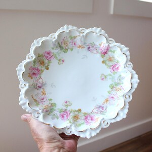 May include: A white porcelain bowl with a scalloped, gold-trimmed edge featuring a delicate floral pattern of pink and yellow roses with green leaves arranged in a circular design around the center of the dish, which has a smooth white finish.