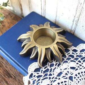 May include: A brass-toned metal candle holder shaped like a sun with radiating rays and a circular center opening. The object features a smooth, metallic finish with darkened recessed areas between the rays, creating a dimensional appearance.