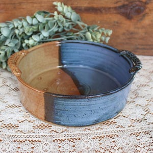 May include: A round ceramic baking dish featuring a two-tone glaze with one half in a warm tan color and the other half in a speckled blue finish. The dish includes two small, textured handles on opposite sides for gripping and lifting.