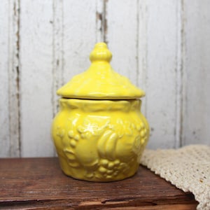 May include: A bright yellow ceramic jar with a lid, featuring embossed floral and fruit designs. The jar sits on a wooden surface, with a white and beige crocheted cloth in the background. The jar is a decorative storage container.