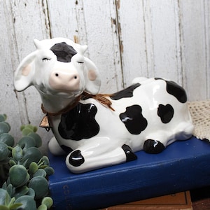 May include: A ceramic cow figurine featuring a glossy white finish with black spots, closed eyes, and small horns. The cow wears a thin brown twine collar adorned with a small, metallic, triangular-shaped bell around its neck.