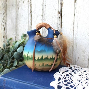 May include: A rounded ceramic vessel with two spouts connected by a handle wrapped in tan leather cord. The surface features a gradient from blue at the top to yellow and brown at the base, with a painted silhouette of a forest and mountains.