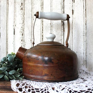 May include: A copper tea kettle with a weathered, dark patina finish. The kettle features a white ceramic handle and a matching white ceramic knob on the lid. It has a short, wide spout and a rounded body, showcasing a metallic construction.