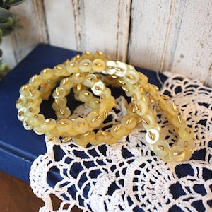 May include: A long strand of circular, pale yellow, translucent buttons linked together with small, metallic gold-toned fasteners. The buttons are arranged in a continuous, flexible chain, creating a decorative trim with a repeating pattern.