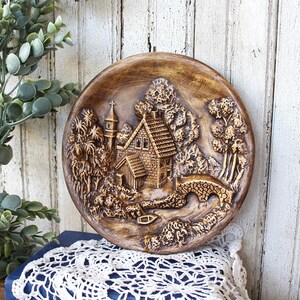 May include: A round brown decorative wall plate featuring a raised relief scene of a house with a steep roof, a tall tower, and a stone bridge over a small stream. The landscape includes textured trees and foliage surrounding the building.