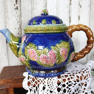 May include: A glossy cobalt blue ceramic teapot featuring a raised relief design of pink flowers and green leaves. The teapot has a brown twisted rope-style handle, a matching lid with a small knob, and a green decorative border around the top.