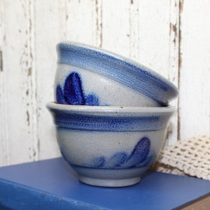 May include: Two stacked ceramic bowls with a speckled light gray finish and a dark blue painted floral pattern. Each bowl features a thick blue band around the rim and a rounded shape with a small base, showcasing a textured, rustic appearance.