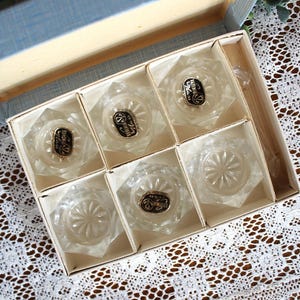 May include: A set of six clear glass coasters with a faceted hexagonal shape and a starburst pattern on the base. Each coaster is housed in a partitioned cardboard box. Several coasters feature a gold and black sticker labeled Bohemia Glass.