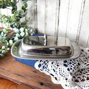 May include: A rectangular silver-toned metal butter dish with a matching lid. The base features a decorative beaded edge, while the lid has a smooth, reflective surface topped with a small, pointed finial handle for easy lifting and placement.