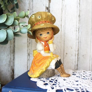 May include: A ceramic figurine of a child wearing a large, wide-brimmed tan hat with yellow polka dots, an orange dress with a white blouse, and brown boots. The figure has blonde hair, blue eyes, and freckles, sitting on a gray base.