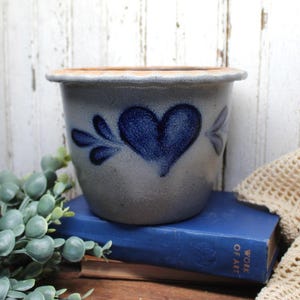 May include: A gray stoneware crock featuring a hand-painted cobalt blue heart design on the front. The vessel has a wide, flared rim with a tan-colored edge and a tapered body. The surface of the ceramic pot has a textured, speckled appearance.