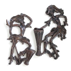 May include: Two bronze-colored wall plaques depicting figures in motion. One figure holds a tall spear next to a drum, while the other figure stands beside a tall, slender drum. Both figures have flowing hair and wear garments with leaf-like details.