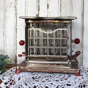 May include: A vintage metal toaster with a rectangular frame, exposed heating coils, and a grid-patterned front panel. The appliance features two round red knobs on the sides, a metallic base with visible oxidation, and a mechanical lever mechanism.