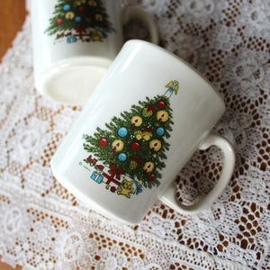 May include: Two white ceramic mugs featuring a colorful illustration of a decorated Christmas tree with red and blue ornaments, lit candles, and wrapped gifts at the base. A small angel figure sits at the top of the tree on each mug.