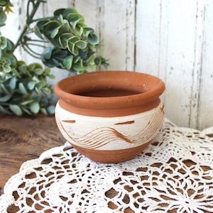 May include: A small, round terracotta pot with a wide, open mouth and a thick, rounded rim. The exterior features a band of white paint around the center, decorated with horizontal, wavy, hand-painted lines that reveal the reddish-brown clay beneath.
