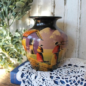 May include: A black ceramic vase featuring a hand-painted landscape scene with a small house, rolling hills, and two figures wearing hats. The artwork uses a palette of purple, orange, yellow, and green tones against a dark background.