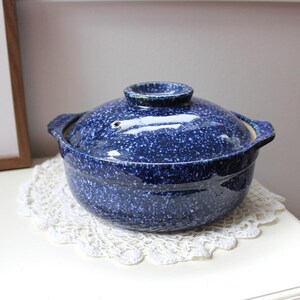 May include: A round ceramic cooking pot with a lid, featuring a deep blue glaze with a white speckled pattern. The pot has two side handles and a central knob on the lid, with a small steam vent hole visible on the top surface of the lid.