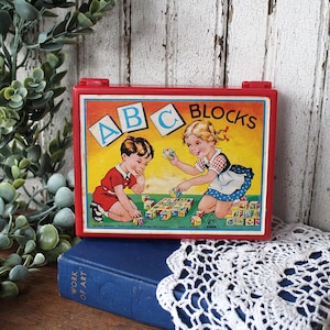May include: A rectangular red plastic case featuring a vintage illustration of a boy and a girl playing with colorful alphabet blocks. The text ABC BLOCKS is printed in black at the top. The case has a hinged lid and a secure latch closure.