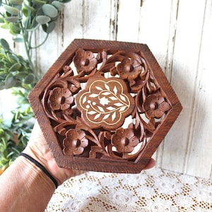 May include: A hexagonal brown wooden trivet featuring intricate hand-carved floral patterns and a central inlay design of white leaf motifs. The openwork carving displays detailed petals and stems throughout the dark wood surface.
