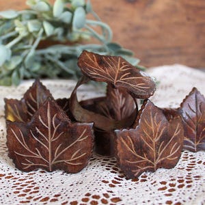 May include: A set of dark brown napkin rings featuring a carved leaf design with visible veins. Each ring is shaped like a single leaf, showcasing a natural wood-like texture and deep brown tones, arranged together to display the detailed leaf patterns.