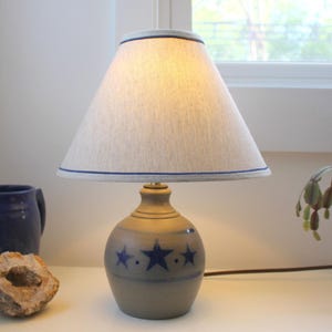 May include: A small, decorative lamp with a beige lampshade trimmed with a blue border. The base is a gray ceramic with blue star designs. The lamp is on a white surface.