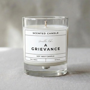 Può includere: Una candela profumata in cera di soia in un bicchiere trasparente. L'etichetta bianca recita "Smells like A GRIEVANCE". La candela è da 100 ml e ha una durata di 24 ore. La cera è bianca.