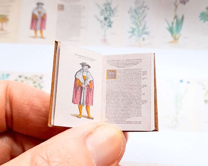 May include: A miniature book with a brown leather cover. The book is open to a page with an illustration of a man in a red and gold robe. The text on the page is in a black font.