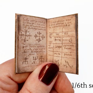 Pu&ograve; includere: Un libro in miniatura con una copertina in pelle marrone e pagine invecchiate. Il libro &egrave; aperto su una pagina con testo scritto a mano e simboli. Il testo include le parole "be semtor etsaluater" e "Con Senna serua". Il libro &egrave; in scala 1/6.