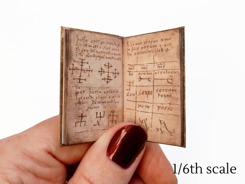 Pu&ograve; includere: Un libro in miniatura con una copertina in pelle marrone e pagine invecchiate. Il libro &egrave; aperto su una pagina con testo scritto a mano e simboli. Il testo include le parole "be semtor etsaluater" e "Con Senna serua". Il libro &egrave; in scala 1/6.