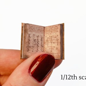 Pu&ograve; includere: Un libro in miniatura con una copertina in pelle marrone e dettagli dorati. Il libro &egrave; aperto su una pagina con testo scritto a mano e simboli. Il libro &egrave; in scala 1/12.