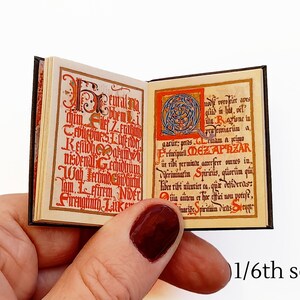 Pu&ograve; includere: Un libro in miniatura con una copertina nera e lettere rosse e dorate. Il libro &egrave; aperto su una pagina con un'illustrazione rossa e dorata e testo in un carattere di stile medievale. Il testo dice "eturalim bhemi gar&uuml;r: pras ille Ragione in preemiari&uuml;n a rmam a kimo Kefidhearum Primiparil. DEZAPRZAR nisdenal Gechidhum ir ribi permintu amerfere omnes in. Vifiriminatin Spiriris, q&uuml;ori&icirc;m qui Wab echinochiiber rihi nuntiet ta, que defideras: Sirrnginth Luk lom Ffirem. NiDELi autem et her offici non poteft, unri&otilde;e Spirituia viens ot".