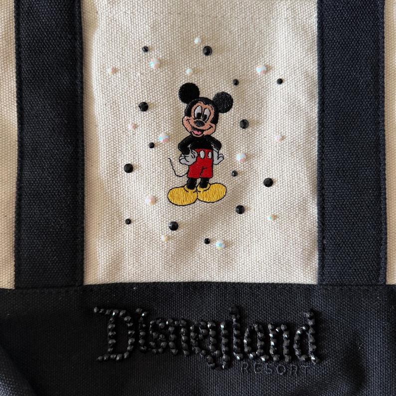 Op de afbeelding: Cr&egrave;mekleurige draagtas met zwarte accenten en een Mickey Mouse-ontwerp. De tas heeft een geborduurde Mickey Mouse, omringd door kleine zwarte en iriserende versieringen. De woorden "Disneyland Resort" zijn uitgeschreven in zwarte strass-steentjes.