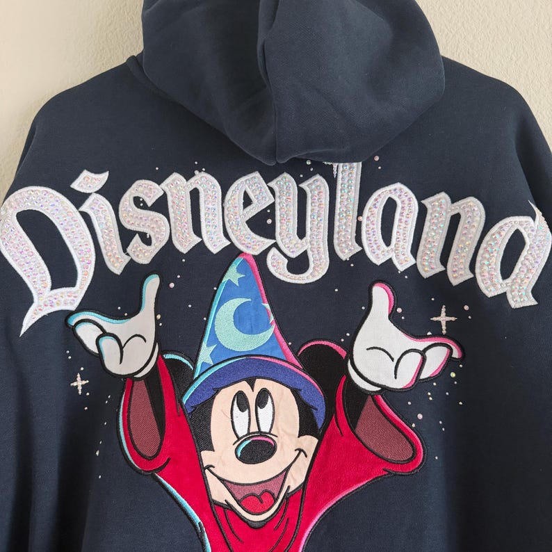 May include: A dark navy blue hooded sweatshirt featuring a large embroidered graphic of Mickey Mouse as a sorcerer with a blue hat and red robe. Above the character, the word Disneyland is written in white letters embellished with clear rhinestones.